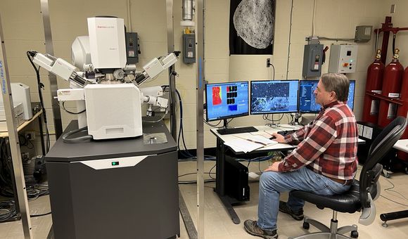 A man sits in front of a large, white machine and a row of computer screens in a lab setting.