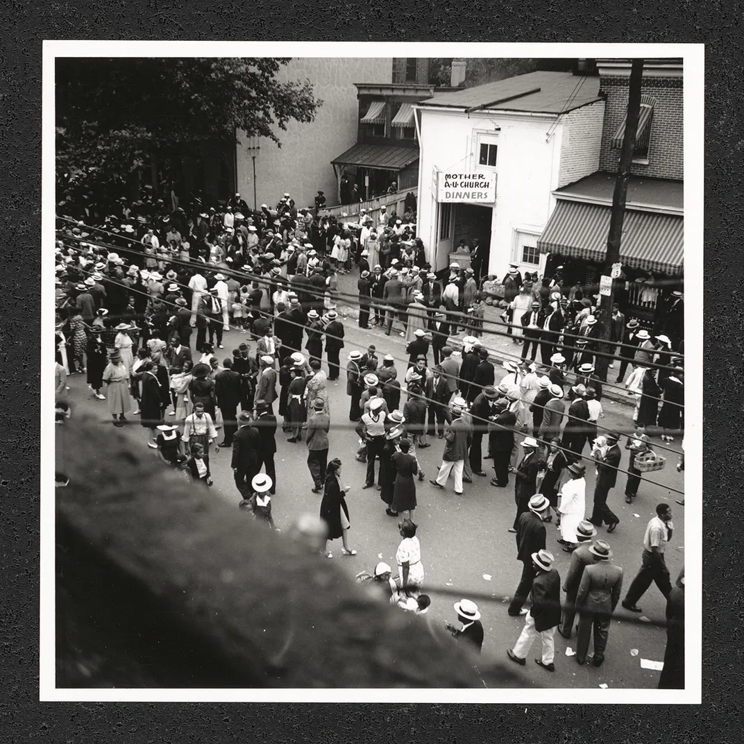 Viewed from a rooftop, a crowd of people process down a city street. Black-and-white photo.