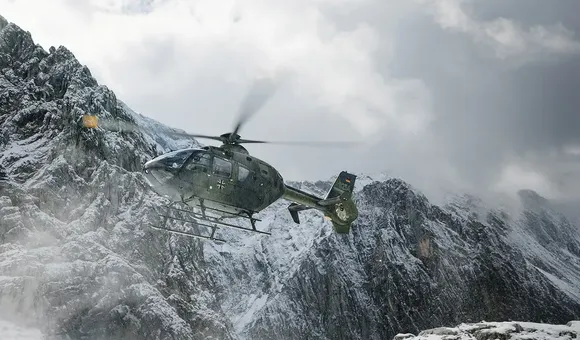 A helicopter flies low over a snow-covered, jagged mountain range. The rotors kick up a light mist of snow against the dark, rocky terrain.