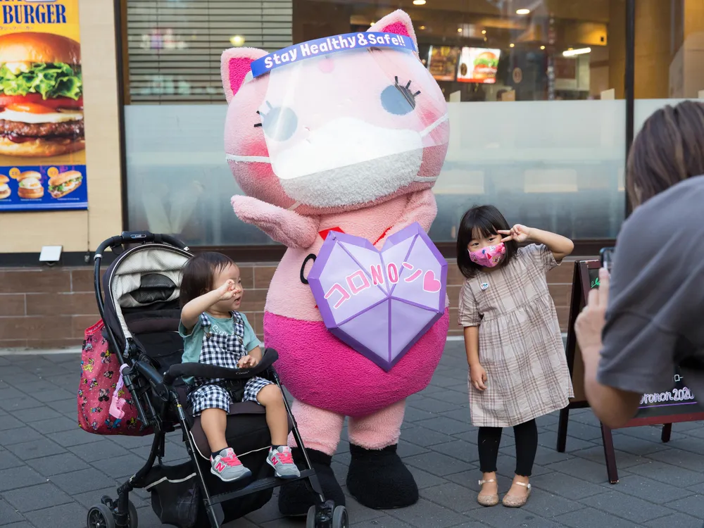 A photo of Japan's Covid-19 awareness Mascot, Koronon, a pink cat with a mask and a purple shield, poses for a photo with two young children. 