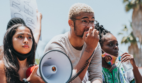 Three young activists are outside at a protest – looking off into the distance. They all appear to be African American. One is holding a bullhorn and has a determined look on her face. Another is holding the bullhorn’s microphone and speaking into it.