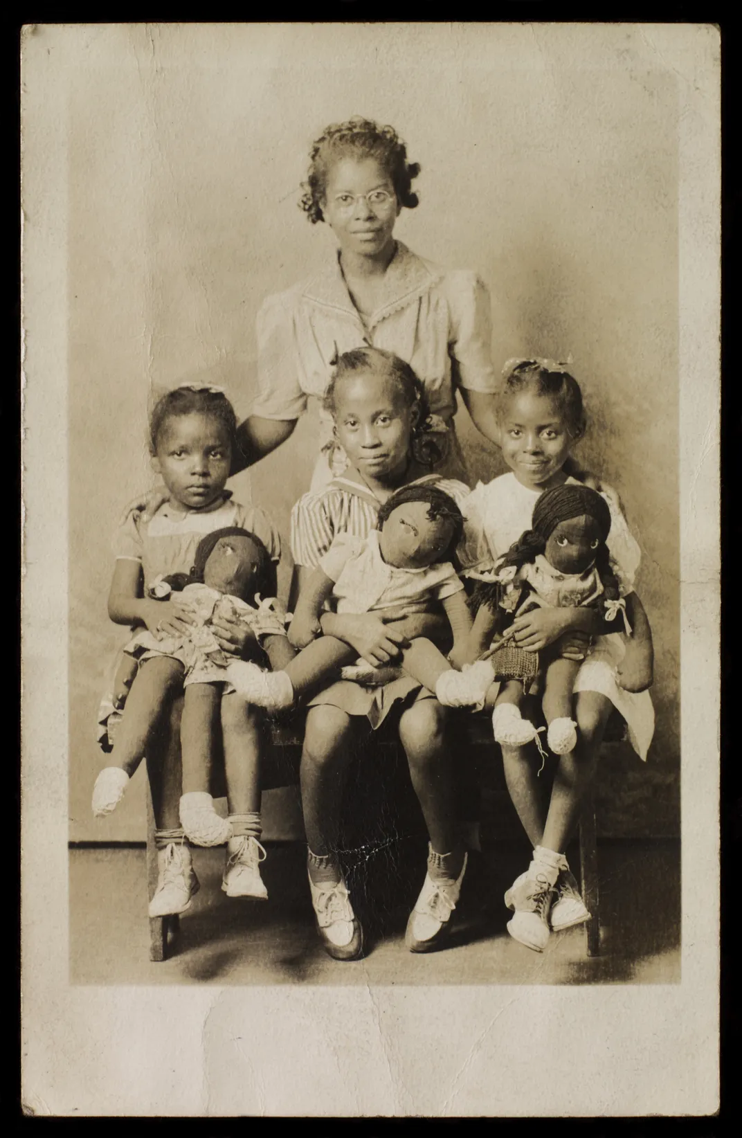 A sepia toned image of one woman standing behind three young girls, seated, each smiling and holding a Black cloth doll in her lap