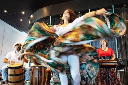 A person dances, turquoise and brown skirt flying and twisting around them, with a drummer and singer performing on the sides.