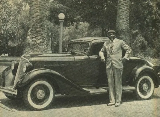 Black and white photograph of a man in a light-colored suit and cap standing in front of a gleaming black car outside with palm trees in the background.