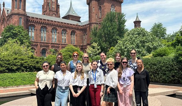 A group of thirteen people stand together in front of green shrubs and the Smithsonian Castle.