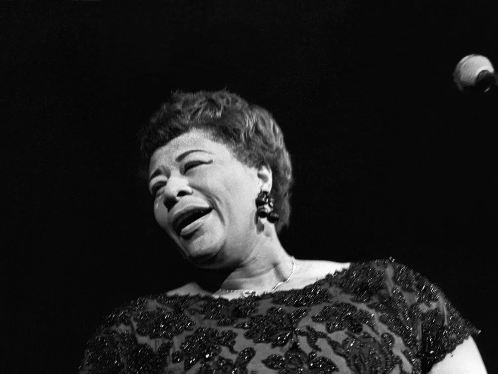 A black and white image, closely cropped to Fitzgerald's profile with the microphone just peeking in on the side; Fitzgerald is singing and turns away from the camera, smiling, mouth half-open and eyes closed, wearing a beaded dress and short cropped hair