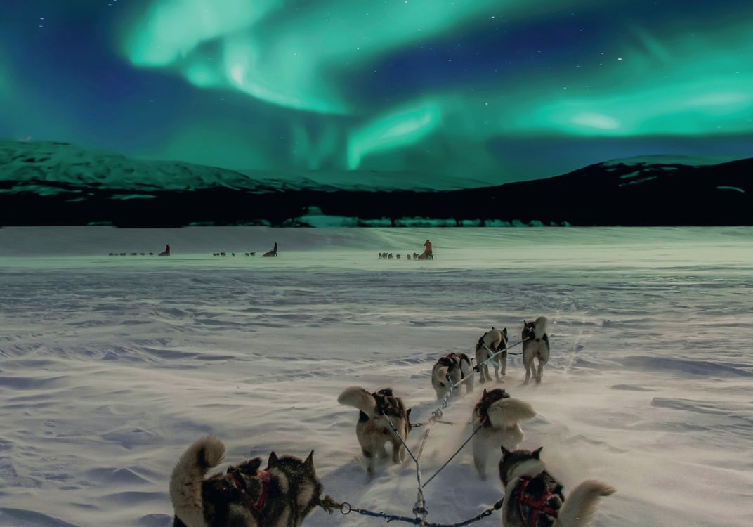 A team of sled dogs pulls a sled across a snow-covered landscape in Arctic Norway as vibrant green Northern Lights swirl across the night sky above distant mountains.