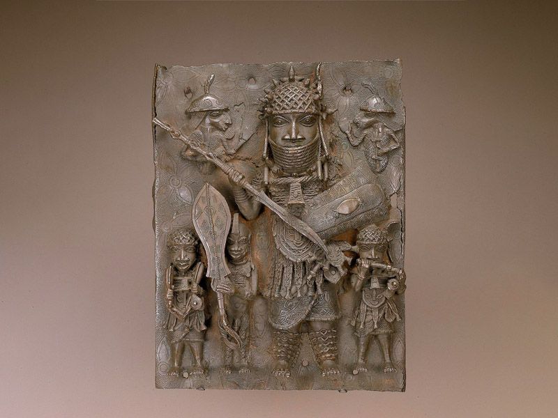 A brown metal plaque that is intricately carved with figures, including a large central figure wearing armor and towering over at least three smaller people standing beneath