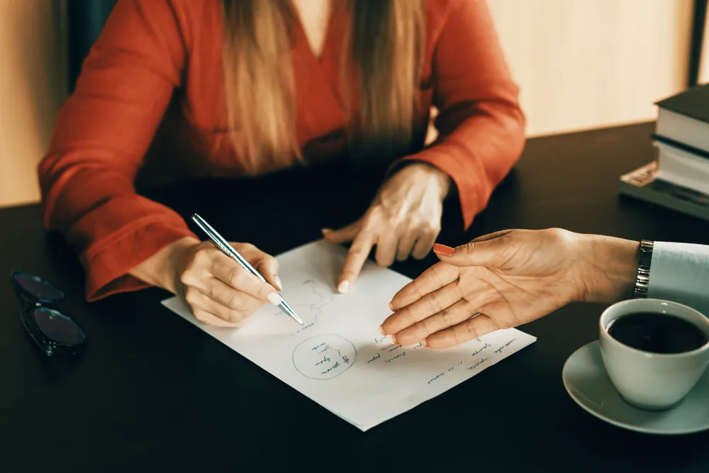 two people at a desk, their faces not visible in the shot, write on and gesture toward a paper with notes