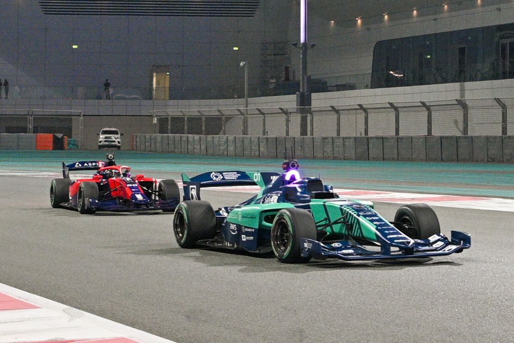 Two driverless race cars, one green (foreground) and one red (background) speed down a straight on the Yas Marina circuit.
