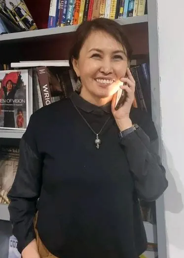 A woman smiles, standing against a bookshelf and holding a cellphone to her ear.