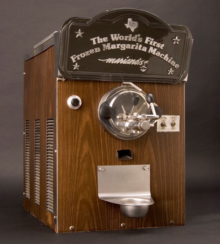 A margarita machine decorated with faux-wood paneling, metal handles and spigots, and a plaque at the top that reads "The World's First Frozen Margarita Machine"