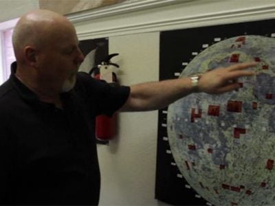 Dennis Hope stands next to a map of the Moon, showing (in red) all the plots of land he’s sold.