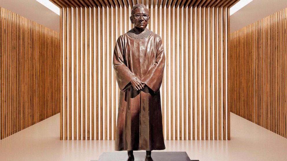 A bronze statue of a short white woman, wearing her Justice robes and standing simply with hands folded in front of her, wearing glasses and her hair in a bun