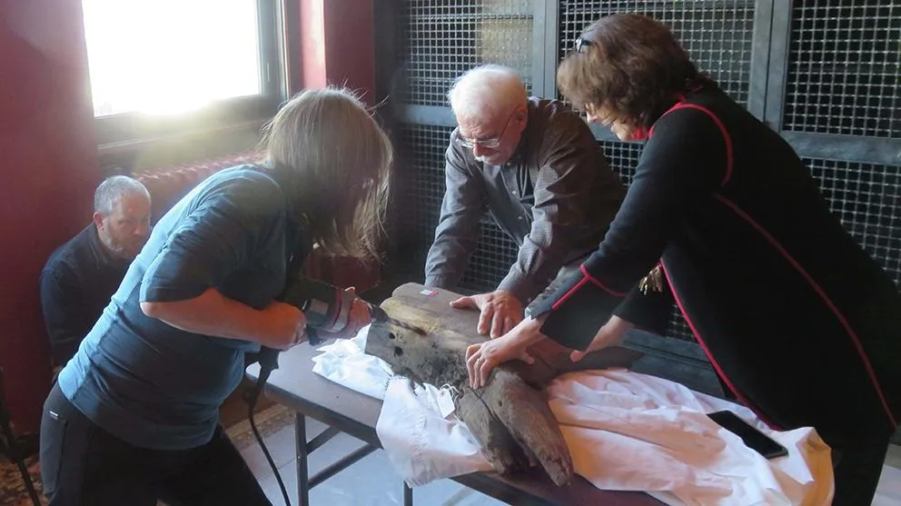 A team of people, including an elderly man in glasses with white hair, stand around a table and look at old fragments of wood