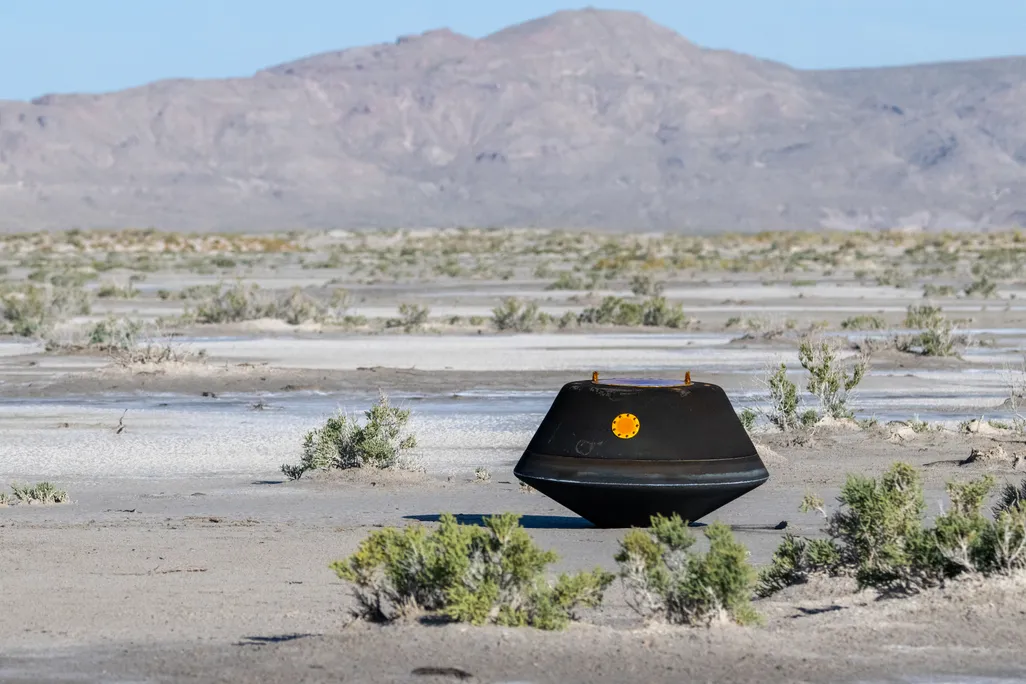 A black, saucer-like capsule sits on the desert surface