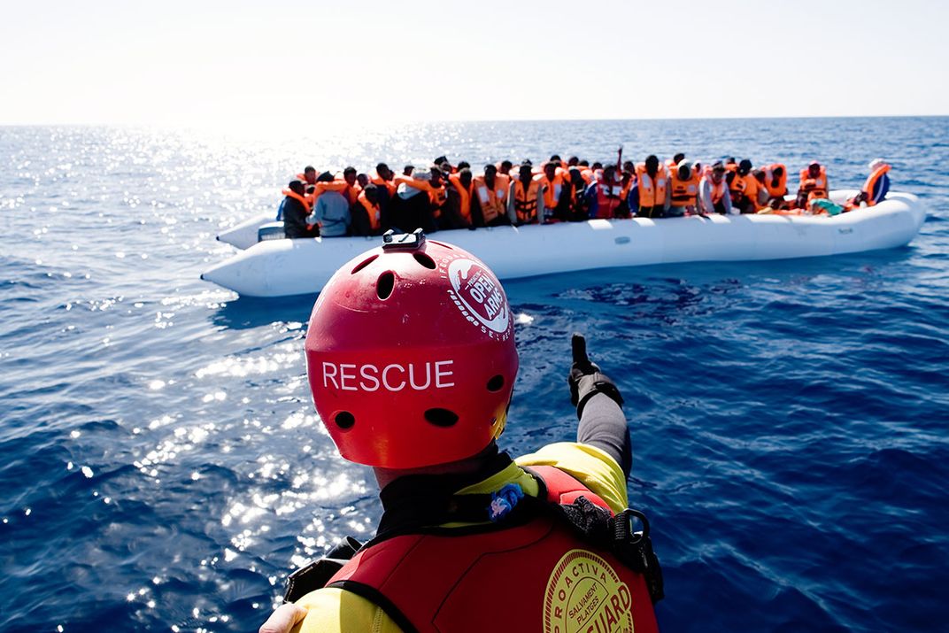 From a boat, a person in safety gear and red helmet that reads RESCUE points toward an inflatable raft full of several dozen people in orange life vests.