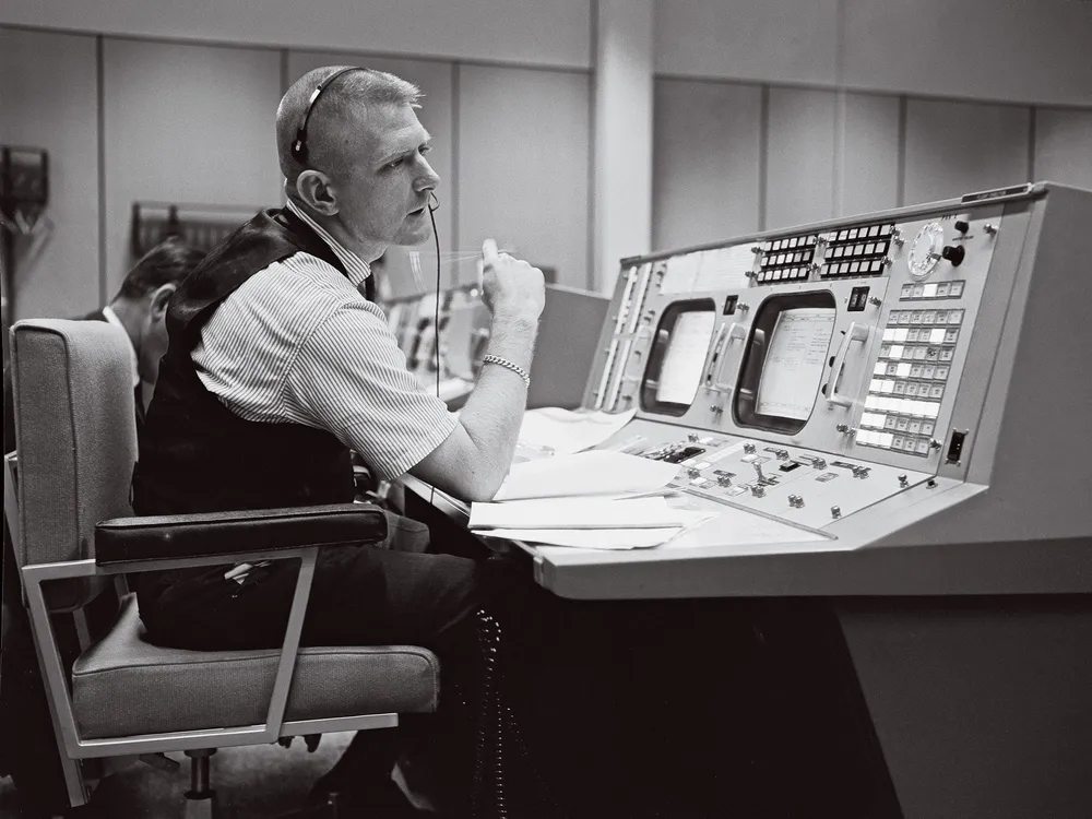 A black-and-white image of a white man in his early 40s sitting at a work console with two display screens and dozens of buttons. The man is wearing a short-sleeve shirt covered by a black vest.