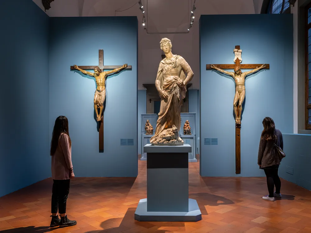 Two people stand in a gallery space with enormous crucifixes on the walls and a large David statue in the center