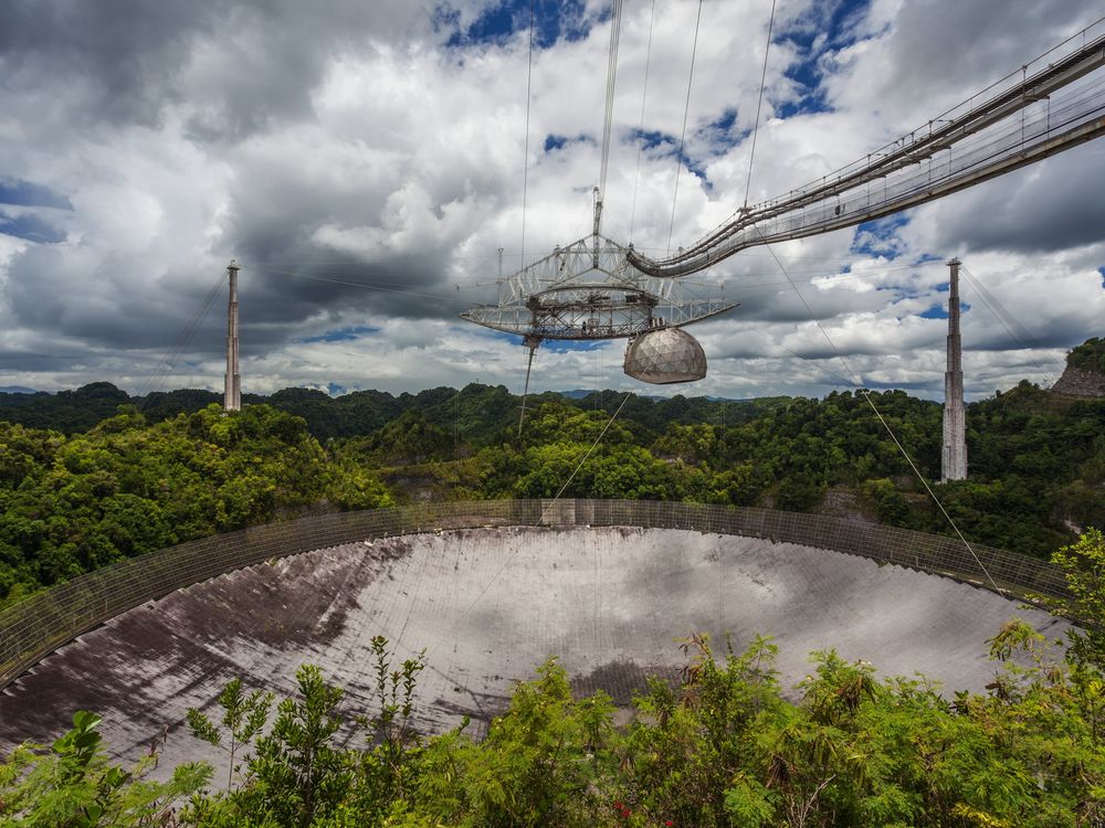 A large metal saucer-shaped object, with a dome attached to its right side, is suspended high above a large dome underneath, the antenna. Behind, an expansive view of blue sky with white clouds and rainforest gives a sense of the telescope's huge size