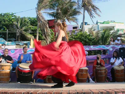 A woman in a long, flowing red skirt, blurry with motion, dances in front of a line of people seated and playing barrel-shaped drums. Palm trees sway in the background.