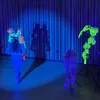 Armando Lopez-Bircann wears a cobalt blue bodysuit and skirt on a stage bathed in blue light. They have a neon green, blue, and pink headdress on and face away from the audience, with in front of their shadow. To the right are props including a neon green