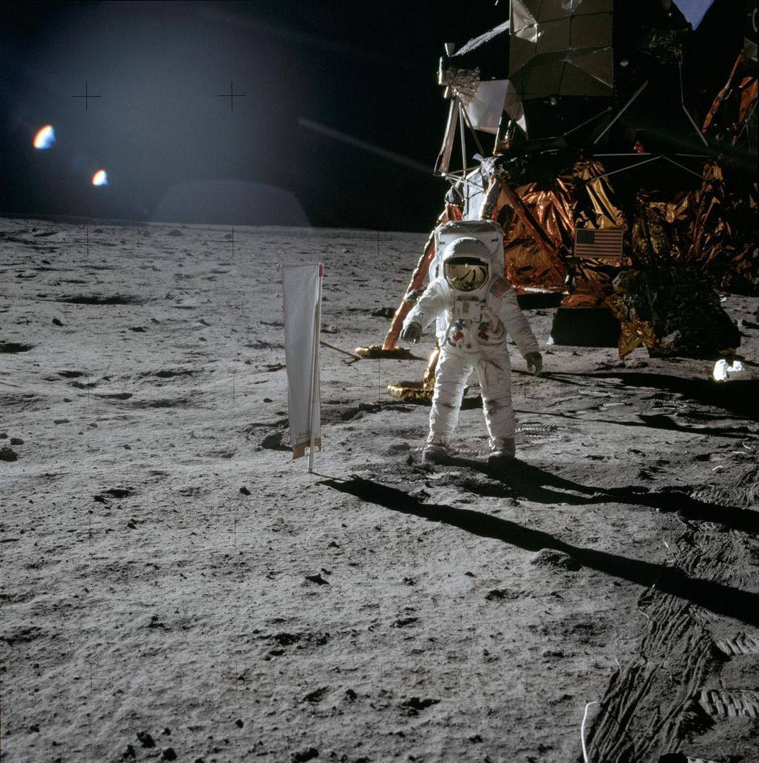 Astronaut Buzz Aldrin stands on the surface of the Moon wearing a white astronaut suit and helmet. Behind him is the lunar module, which bears an American flag and appears as if wrapped in shiny foil.