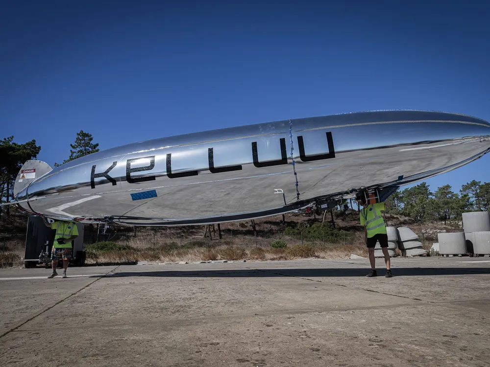 Two people standing outdoors on a paved surface holding a long, silver, cigar-shaped airship drone that features the text "KELLUU" in large black letters along its side.