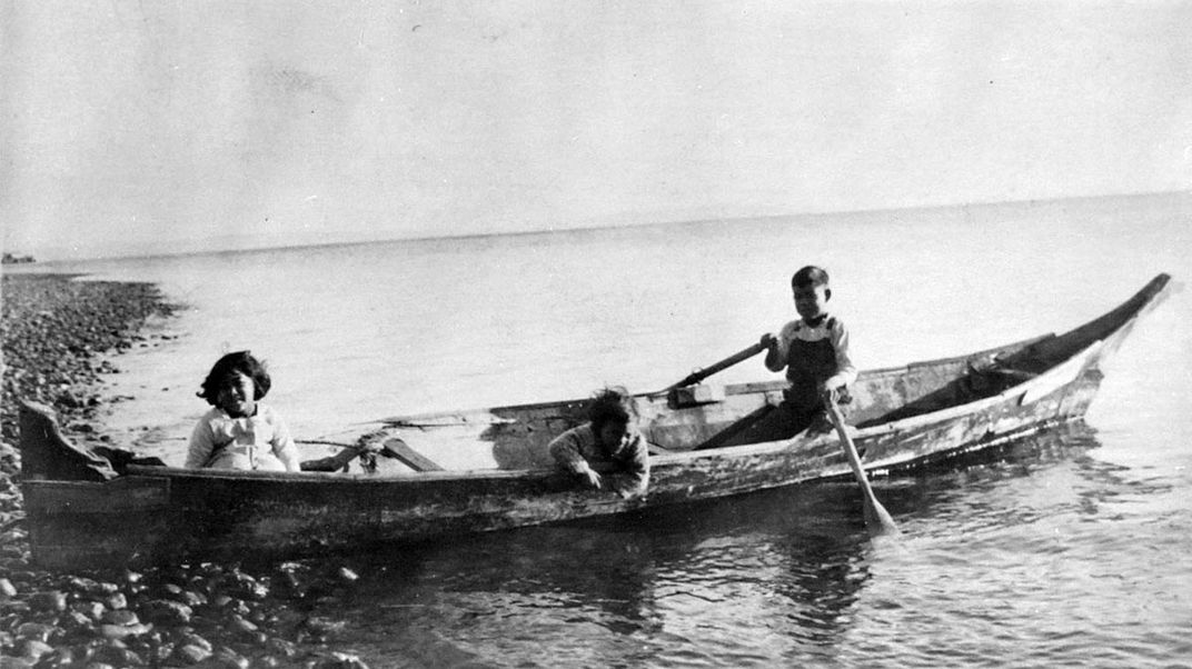 Three young kids sit in a canoe docked at the edge of a rocky shore. One child holds two oars in the water. Black-and-white archival photo.