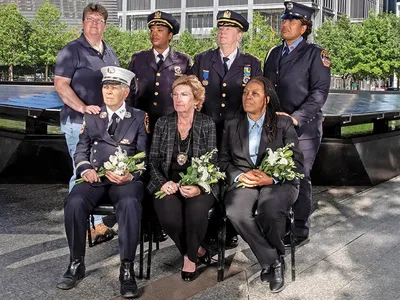 group of women first responders in uniform, standing and sitting with Ground Zero in the background. Three are seated and holding white bouquets