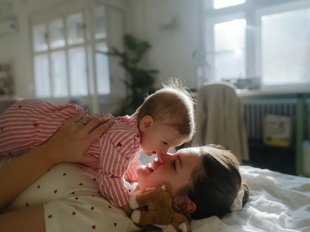 a woman lies on a bed holding her baby to her face, touching noses