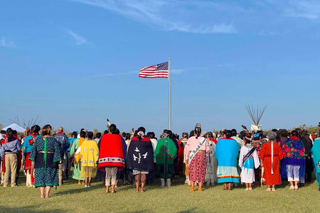 Members of the Kiowa Gourd Clan Ceremony stand as the flag of Spencer “Corky” Sahmaunt is raised. Carnegie, Oklahoma; July 4, 2019. Mr. Sahmaunt served in the U.S. Army during the Korean War and was a member of the Kiowa Black Leggings Warrior Society, as well as the Kiowa Gourd Clan.The Kiowa Flag Song, analogous to the Star Spangled Banner, accompanied the flag-raising. (Photo courtesy of Mari Frances Sahmaunt, used with permission)