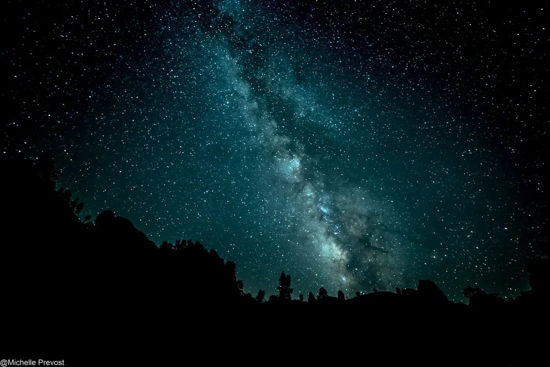 the milky way appears blue over some dark trees