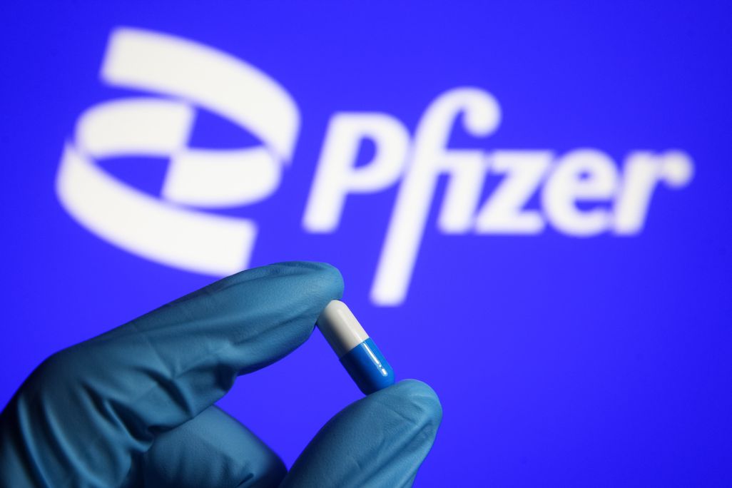A gloved hand holds a pill in front of the Pfizer logo