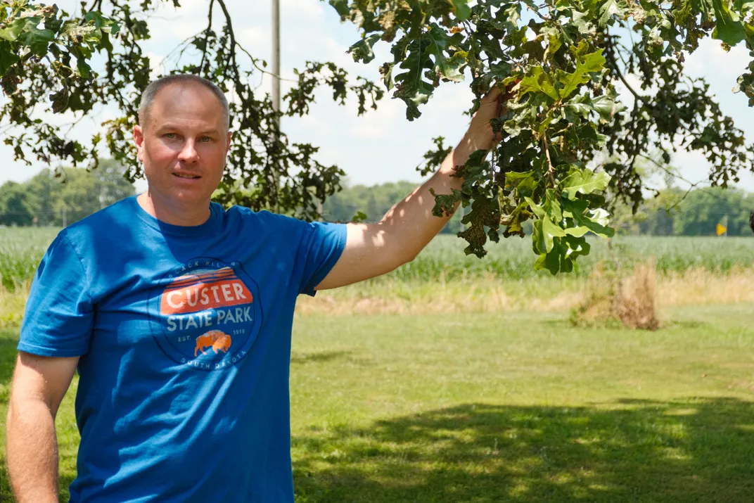 a man wearing a blue Custer State Park shirt holds onto a tree branch and faces the camera