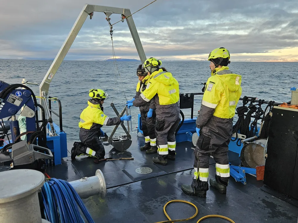 researchers in yellow hats and coats stand—one kneels—on the deck of a small research vessel on the ocean, examining a sediment core