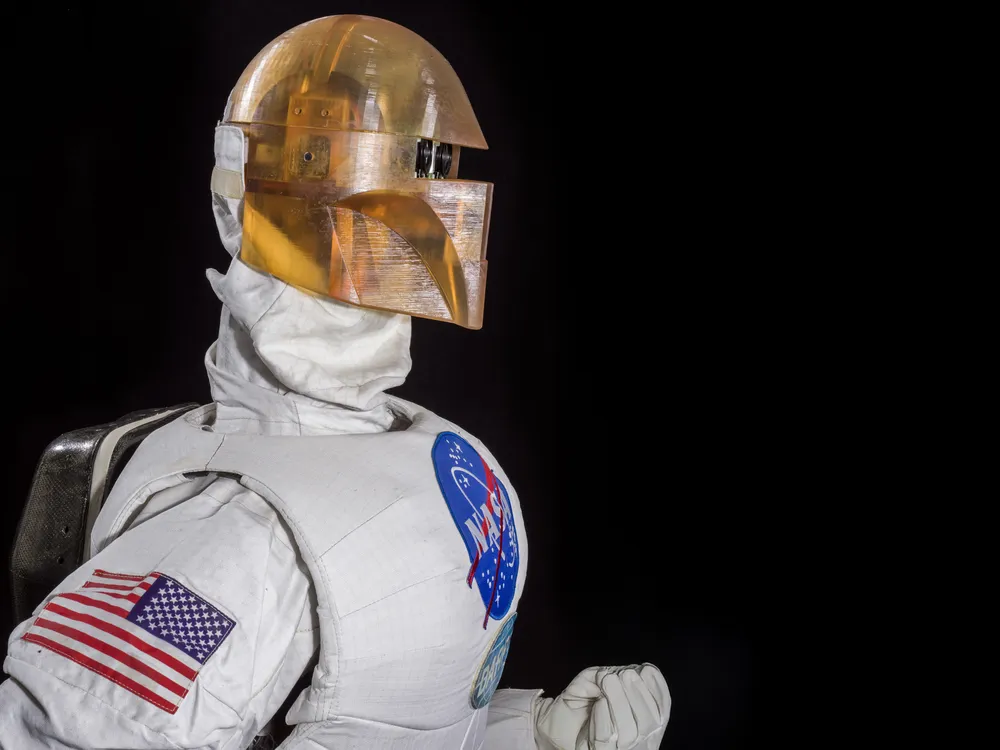 Photographed against a black background is a robot in profile wearing a silver uniform and golden-colored helmet.