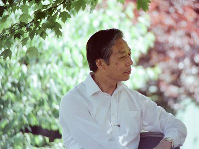 A man in white dress sits with a thoughtful expression on his face outdoors, green and red foliage out of focus behind him. Old color film photo.