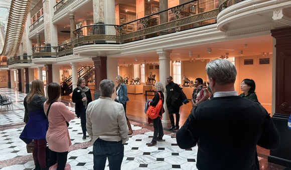 People stand and listen to a speaker in a brightly lit museum room underneath a large gold art installation.