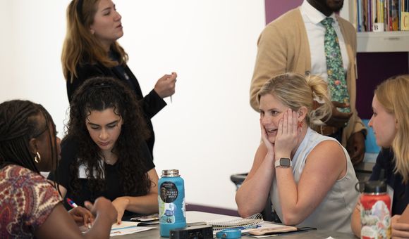 Teachers converse at a table, one woman with blond hair has her hands to her face