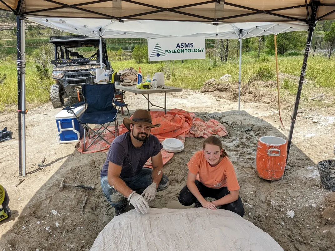 under a tent canopy, a man and a teenage girl squat and smile at the camera in front of the fossil, a white blob, with plaster on it