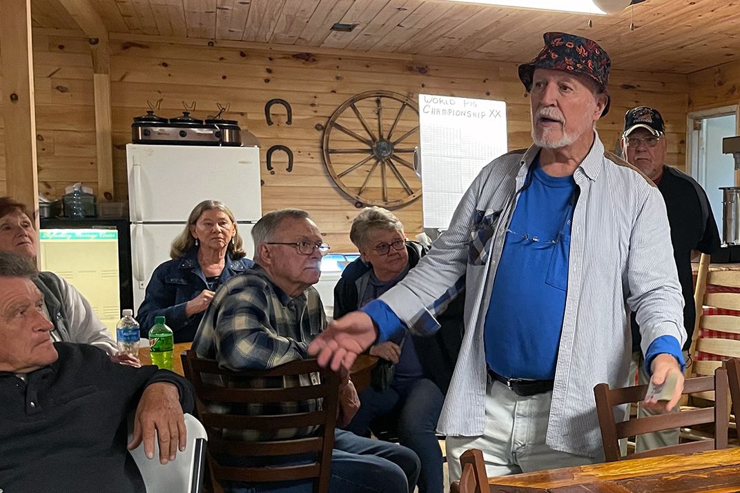 A man in a floral bucket hat stands, speaking to a group of seated people.