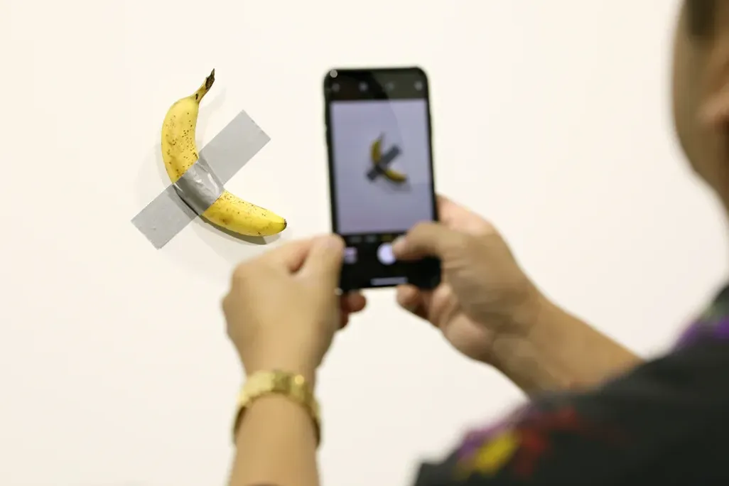 Person taking a photo of a banana taped to a white wall