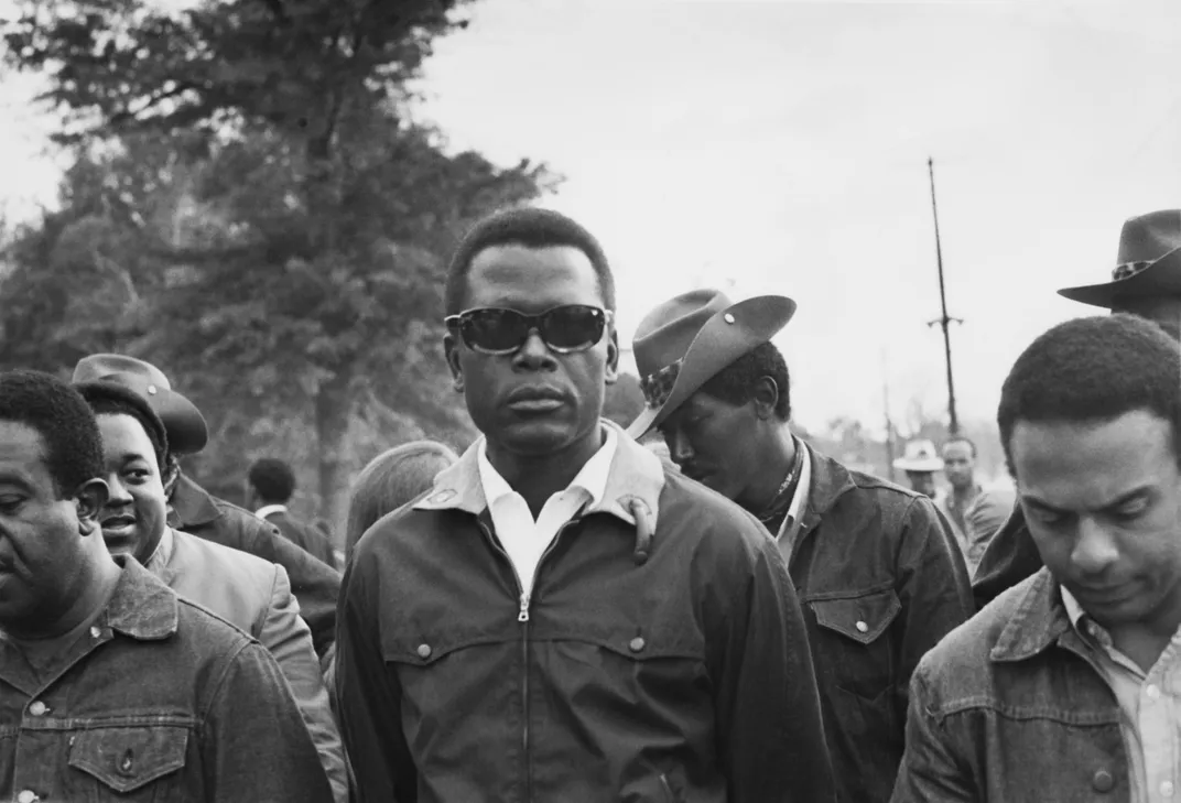 Poitier, center of a crowd of Black men marching, wears a jacket and dark sunglasses and stares you straight in the eye