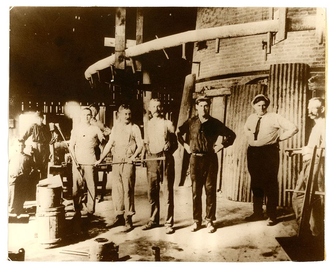 A group of men pose in a row inside a factory room, some holding long metal pipes.