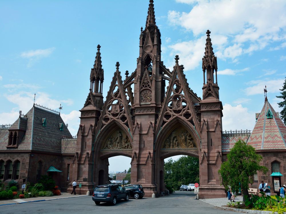 The Gothic revival entrance of Green-Wood Cemetery, two tall pointed archways and a third tall spire in the middle, in a reddish brick color with a blue sky behind