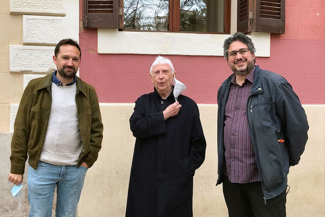 Three  men in coats pose outside, holding or removing their face masks.