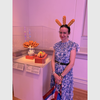 Sarah Rogers Morris wearing a headband with three corndogs poking out stands behind an exhibition case with a vase containing corndogs and a ceramic hot dog.
