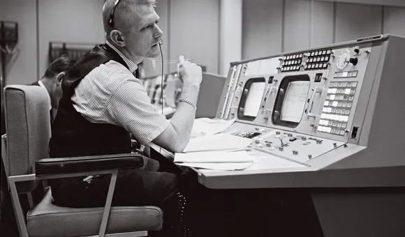 A black-and-white image of a white man in his early 40s sitting at a work console with two display screens and dozens of buttons. The man is wearing a short-sleeve shirt covered by a black vest.