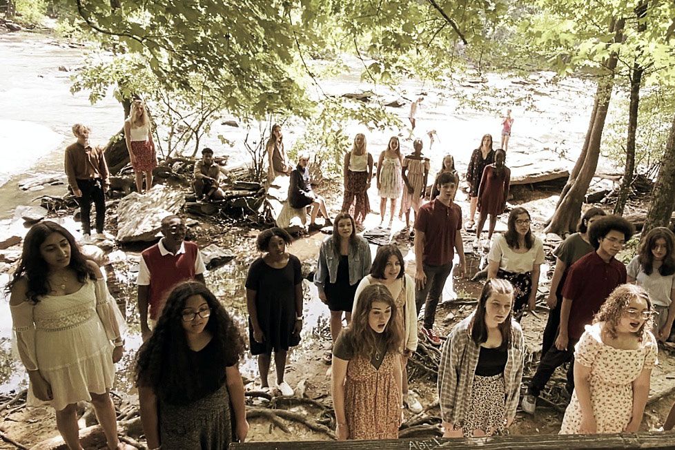 Choir of high school students sing outdoors in dappled sunlight.